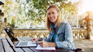 Young woman smiling to the camera while sitting with notepad and laptop computer in beautiful city park, happy Caucasian female posing during work on portable net-book outdoors in warm spring day